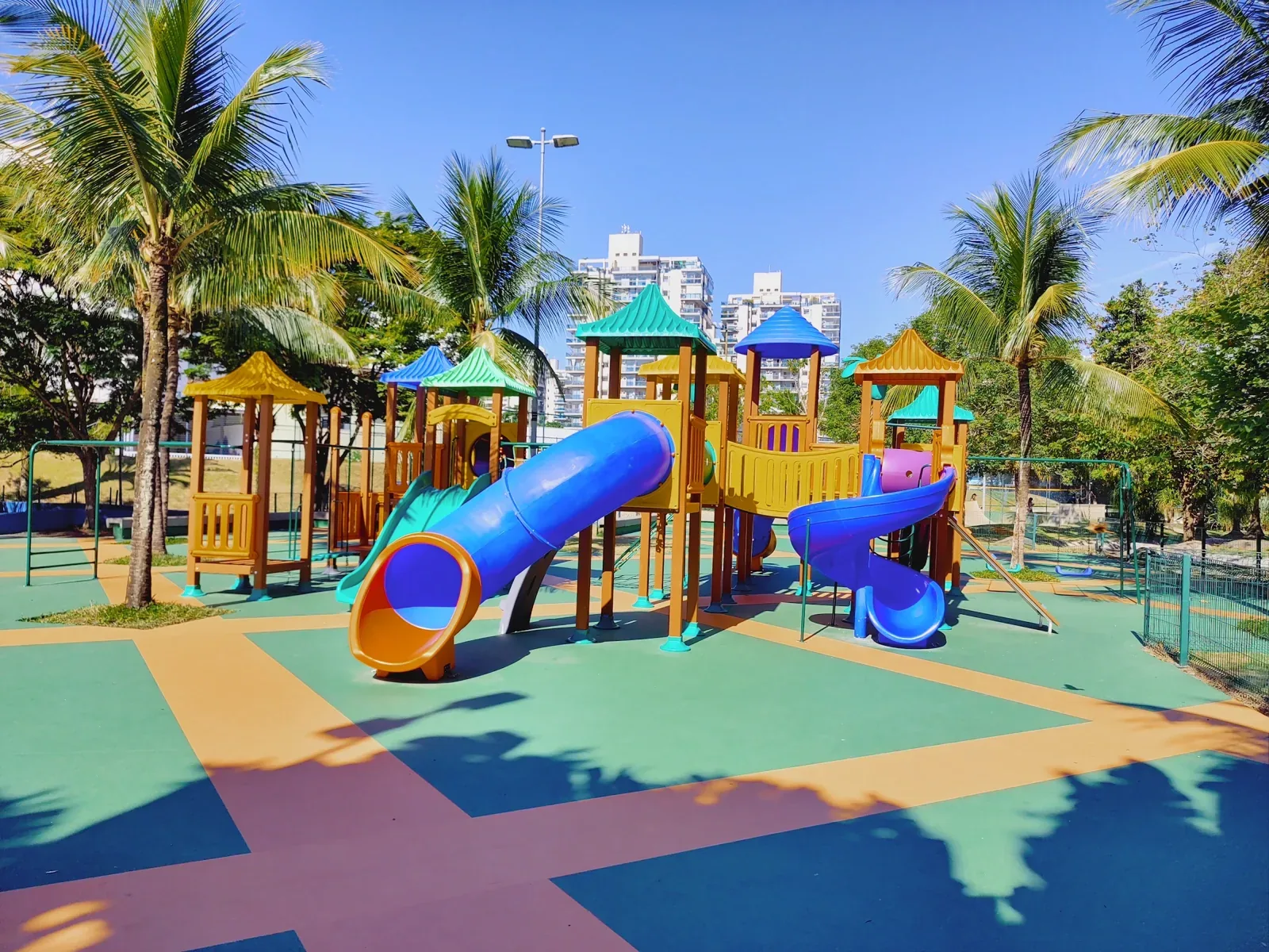 Playground grande com escorregadores em tubo sobre piso emborrachado colorido, entre palmeiras, na Associação Rio 2, Barra Olímpica
